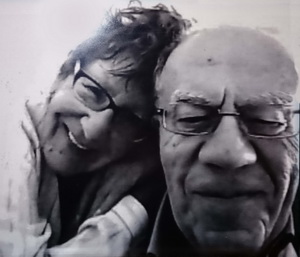 Black-and-white close-up photo of Albert and Jeanine Samaha. They are both older people and each wear spectacles. Jeanine leans on Albert's shoulder. They are both smiling. She is looking directly at the camera, while Albert is looking down as he focuses on something in front of him.
