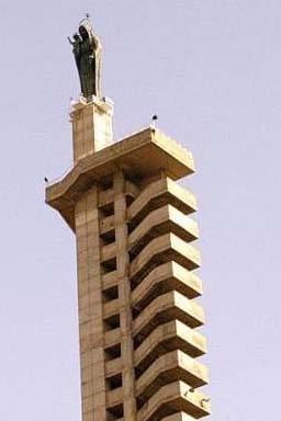 View from the ground of a grey statue of the Virgin atop a tall sandstone-coloured building